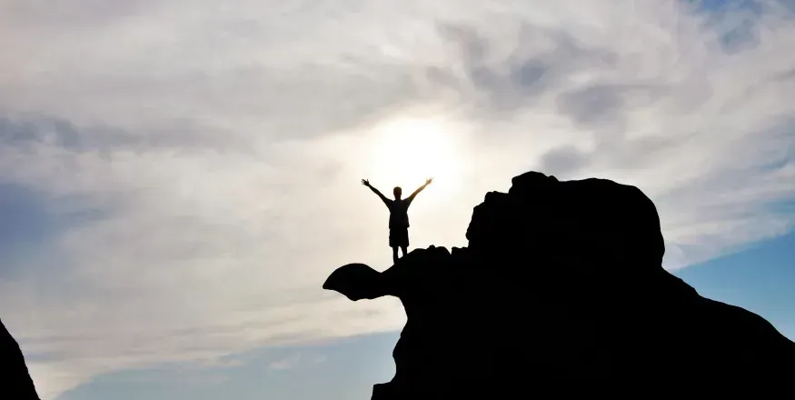 Silhouetted climber standing on a rocky peak with arms raised at sunrise or sunset