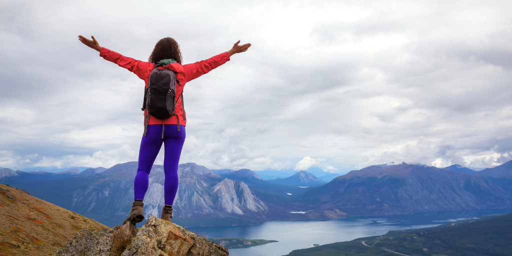 Person in red jacket and purple pants stands with arms raised on a mountain overlook above a lake and peaks