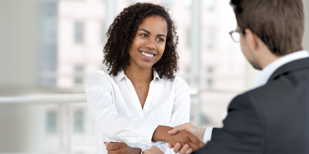 Two businesspeople shaking hands in a bright office, one smiling in a white blouse