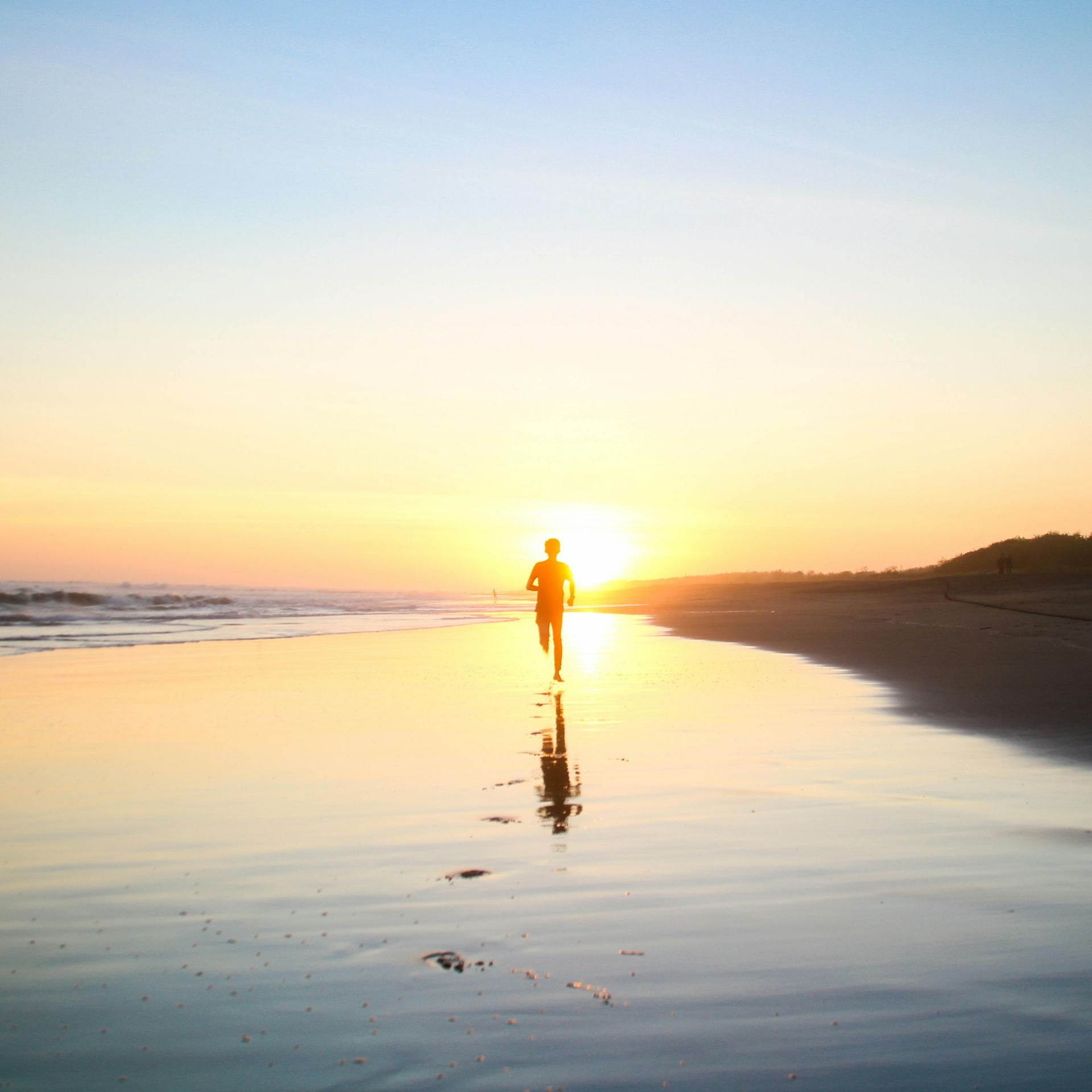 Person running on a beach at sunrise, silhouetted against the bright sun, reflected on wet sand