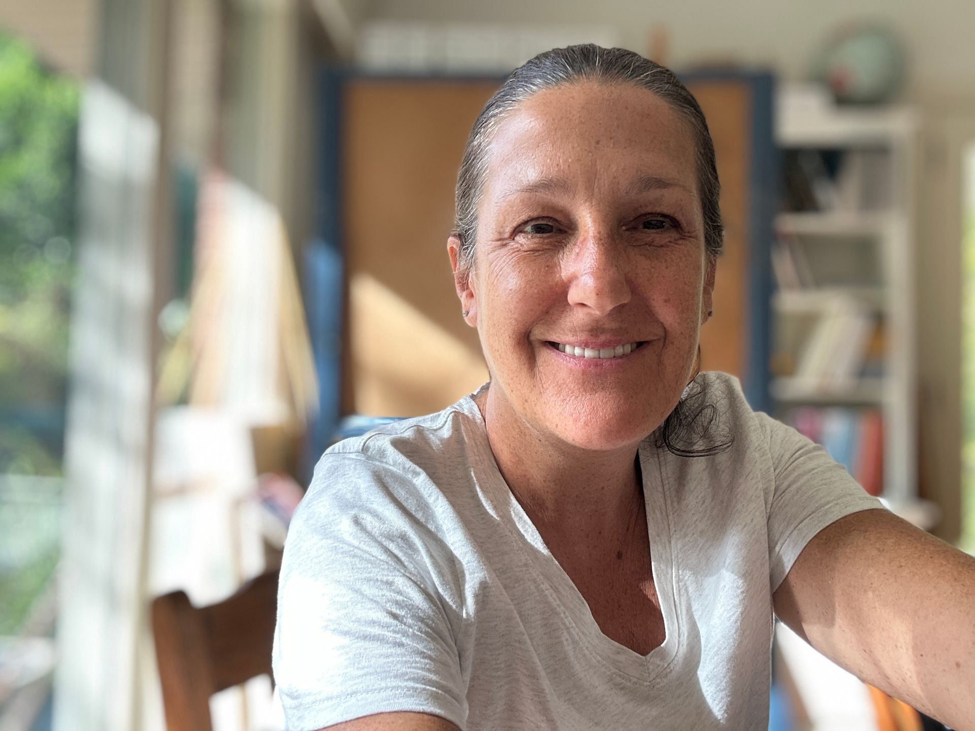 Smiling person in a light gray shirt, leaning toward the camera in a bright home office.