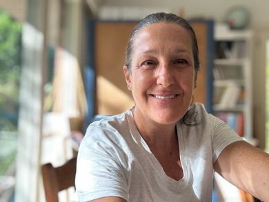 Smiling person in a light gray shirt sitting in a bright room with shelves and a window in the background