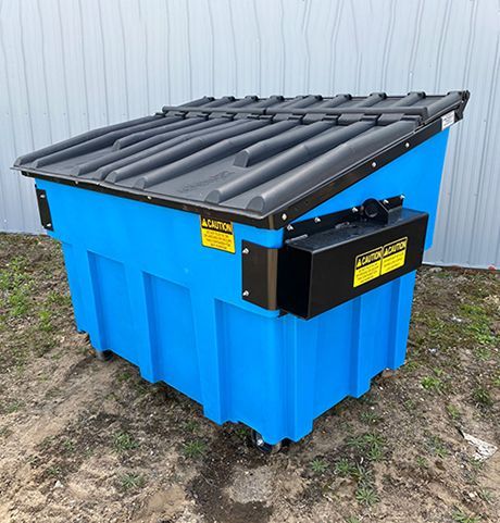 A blue dumpster with a black lid is sitting in the dirt.