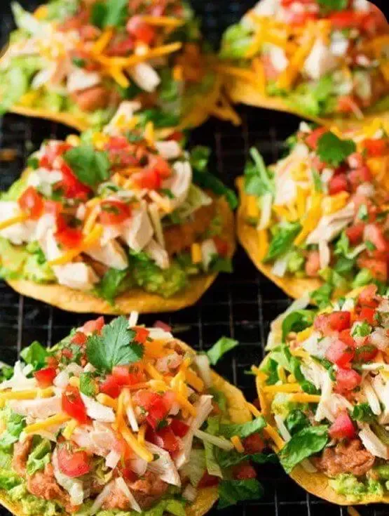 A close up of a bunch of tostadas with guacamole , chicken , tomatoes , cheese and other toppings