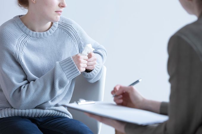 Una persona con un maglione grigio parla durante una seduta di consulenza, mentre un ascoltatore seduto prende appunti.