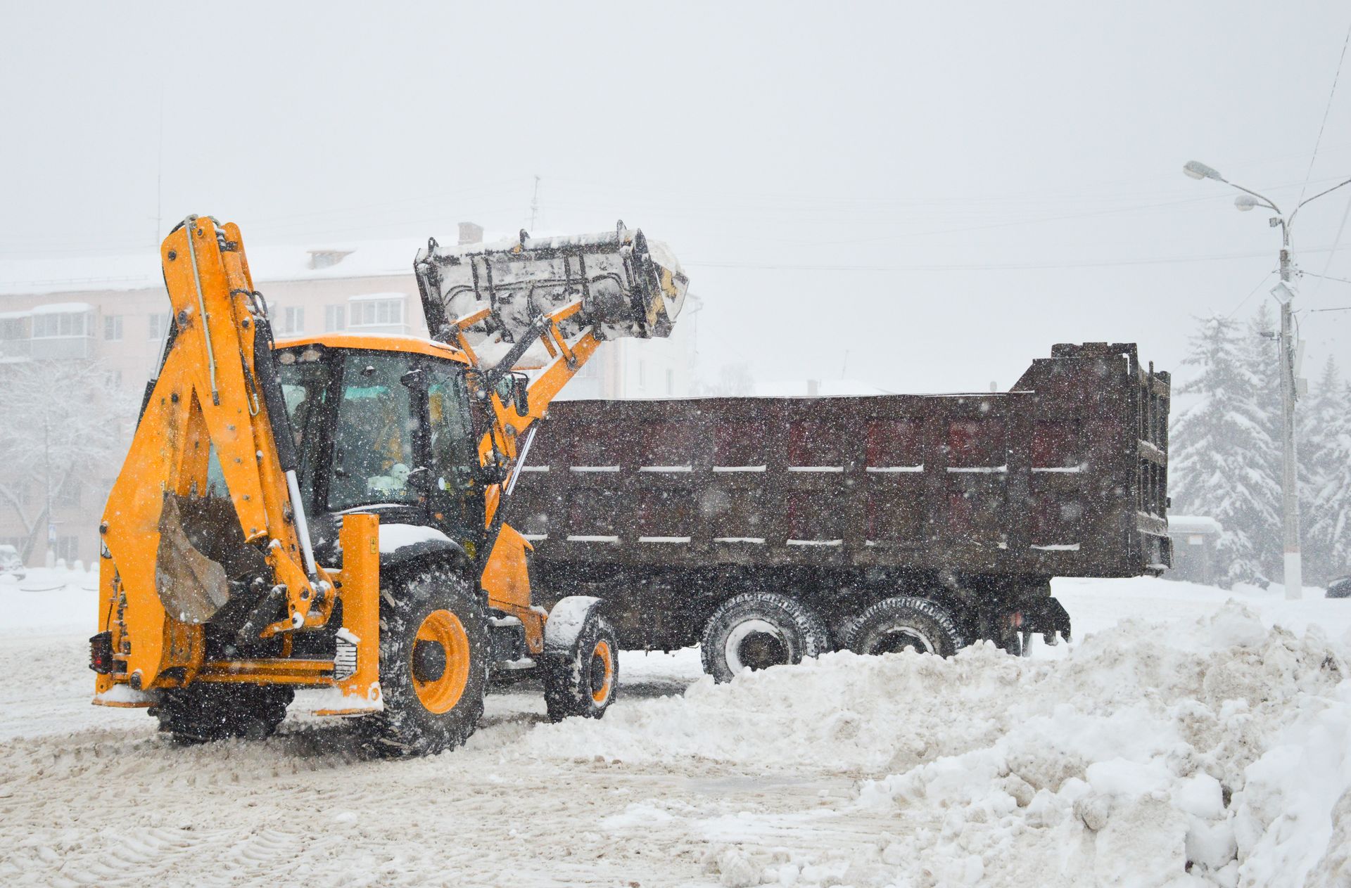 Une pelleteuse jaune charge de la neige dans un camion-benne marron sur une rue enneigée pendant une chute de neige.