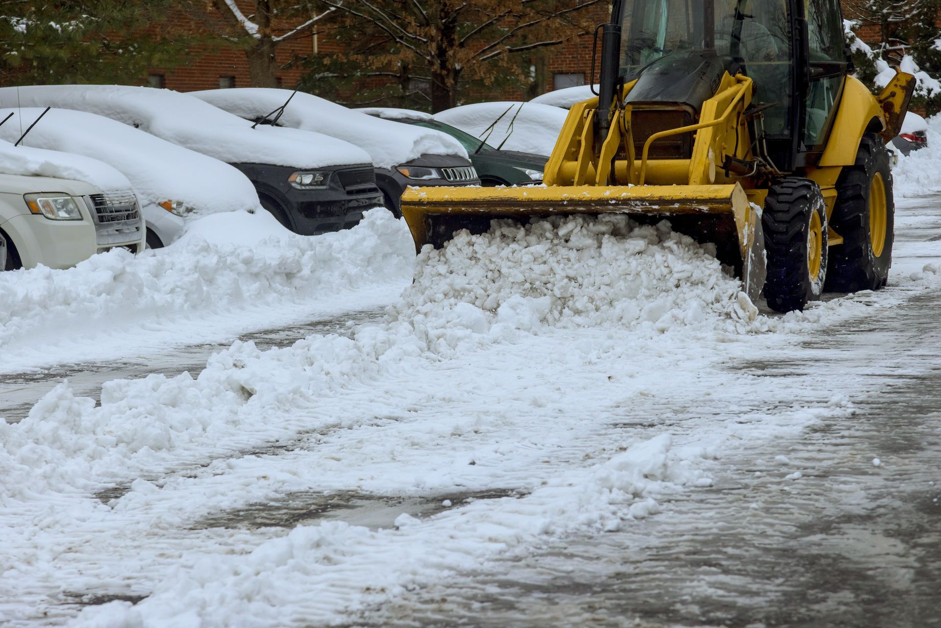 Un tracteur jaune déneige un parking, près de voitures recouvertes de neige.