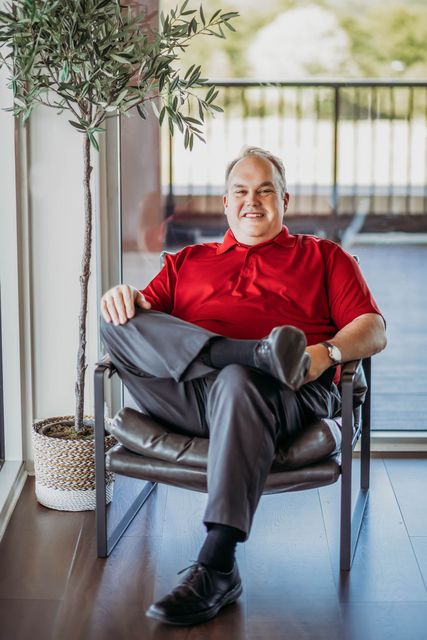 a man in a red shirt is sitting in a chair with his legs crossed .