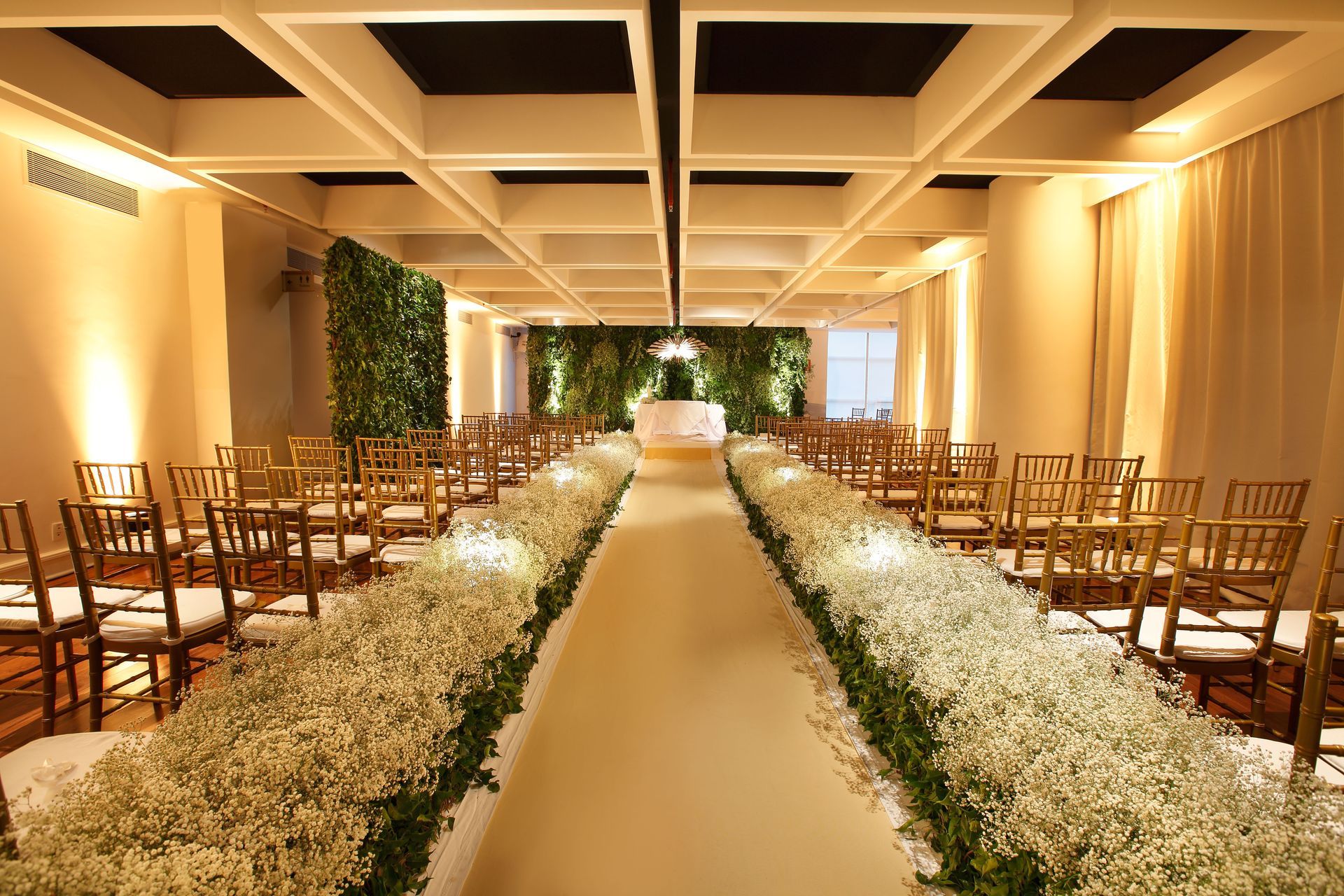 Elegante corredor interno para casamento com flores brancas, cadeiras de madeira e iluminação aconchegante no teto.