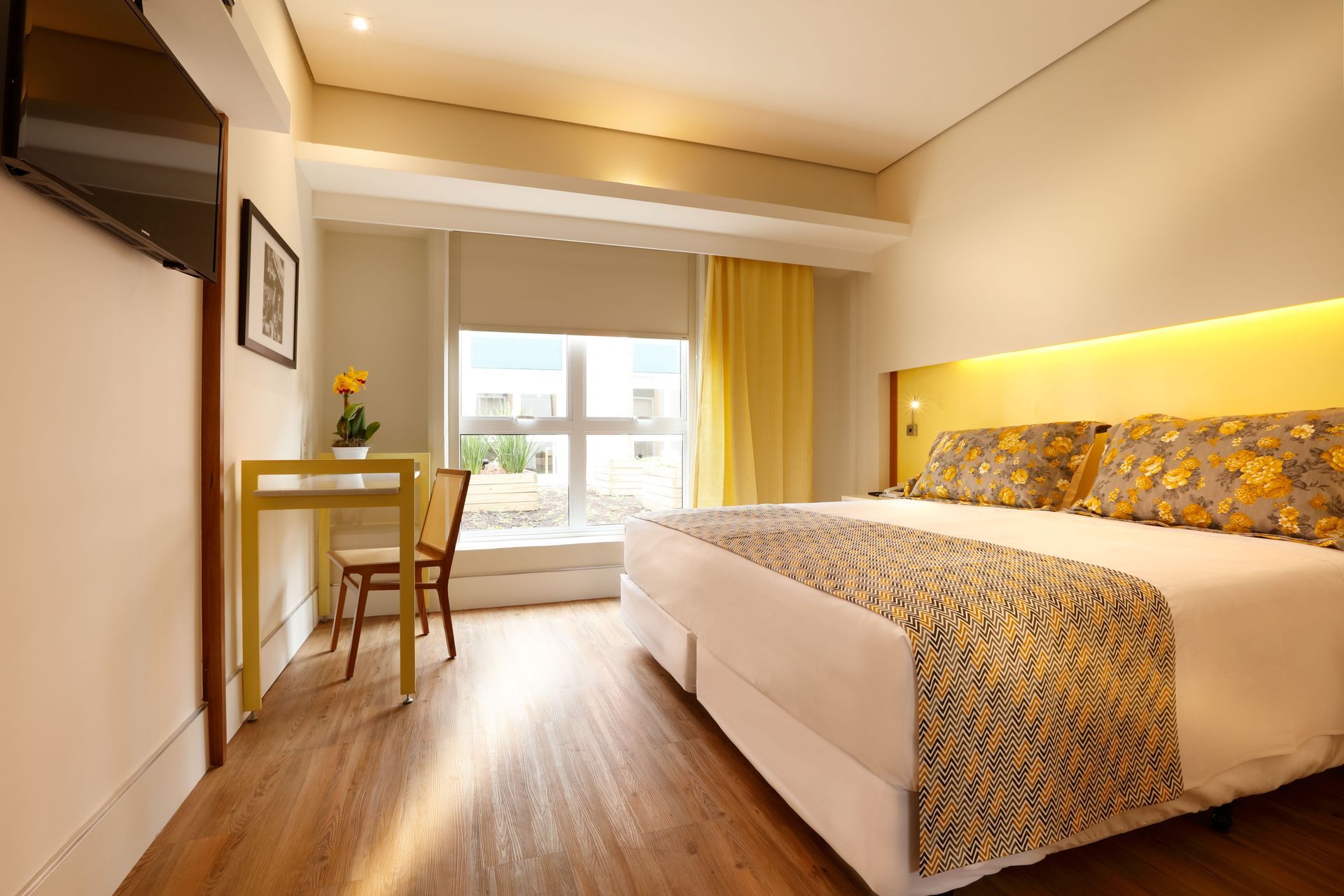 Quarto de hotel moderno com uma cama grande, escrivaninha junto à janela e iluminação amarela aconchegante.