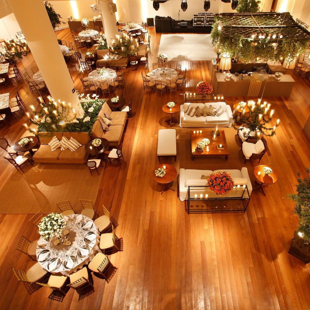 Ambiente elegante para eventos em espaço interno, com piso de madeira, mesas redondas, sofás brancos e iluminação aconchegante com cordões de luz.