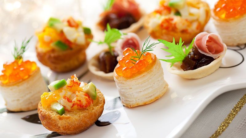 Aperitivos variados em porções individuais, decorados com laranja, servidos em uma travessa branca.