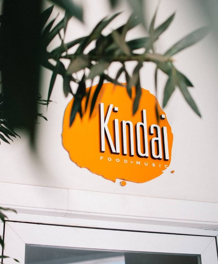 Logotipo laranja da Kindal em uma parede, parcialmente emoldurado por folhas verdes acima.
