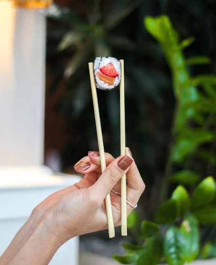 Mão segurando hashis com um rolinho de sushi coberto com salmão e abacate contra um fundo verde desfocado.