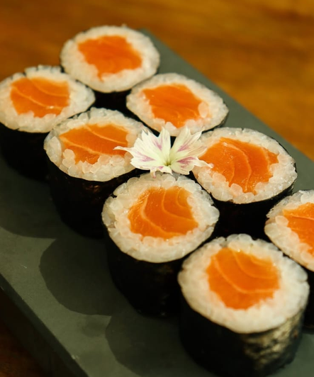 Rolinhos de sushi com salmão alaranjado em um prato escuro, decorados com uma flor branca.