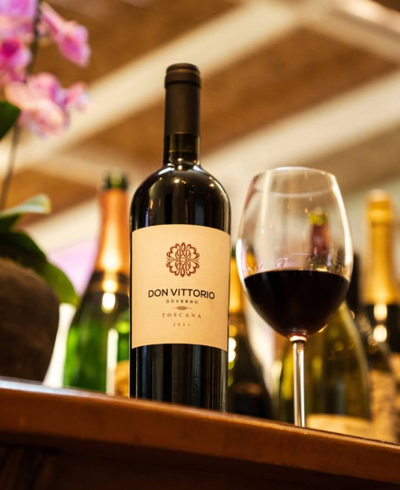 Garrafa de vinho Don Vittorio ao lado de uma taça de vinho tinto sobre uma mesa de madeira, com garrafas desfocadas ao fundo.
