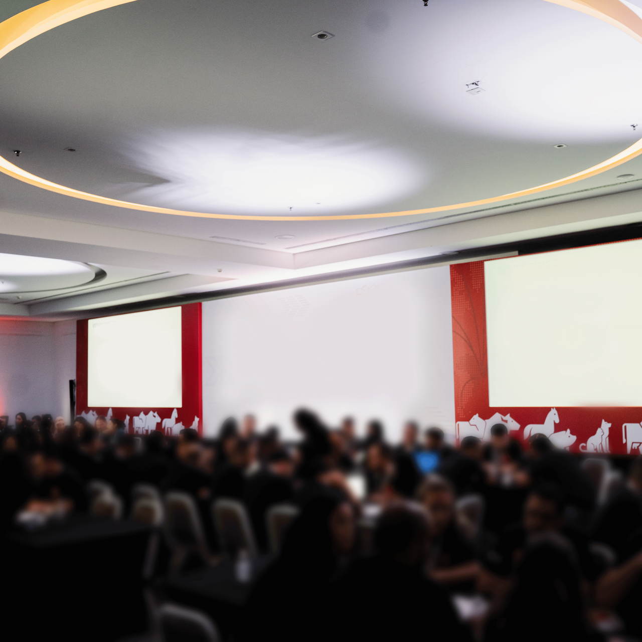 Público sentado em uma sala de conferências de frente para um palco com painéis de parede vermelhos e brancos.