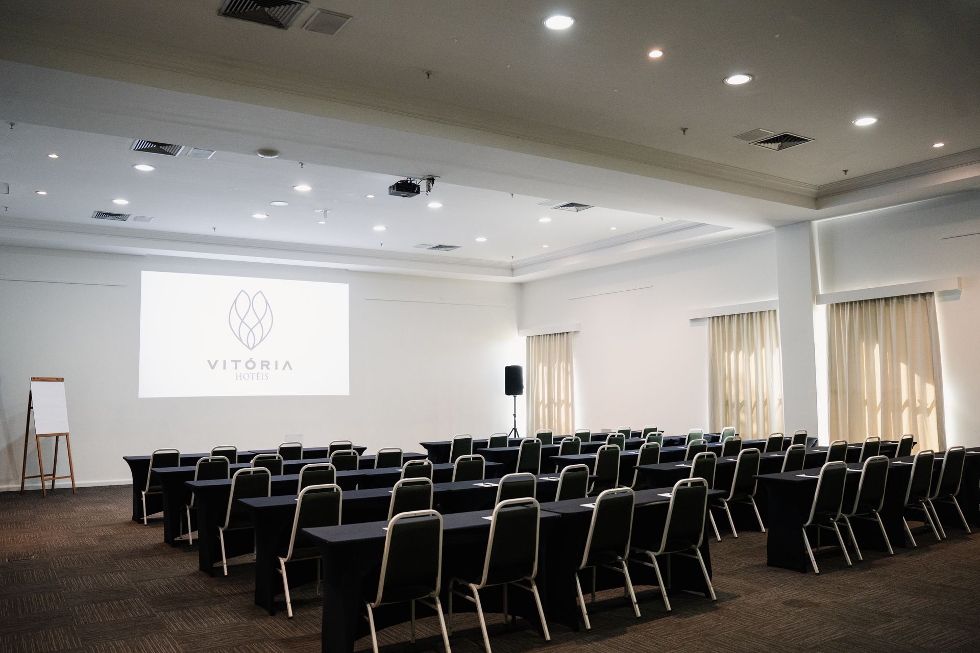 Sala de conferências com fileiras de cadeiras pretas voltadas para uma tela de projeção e um palco, iluminada por luzes de teto.