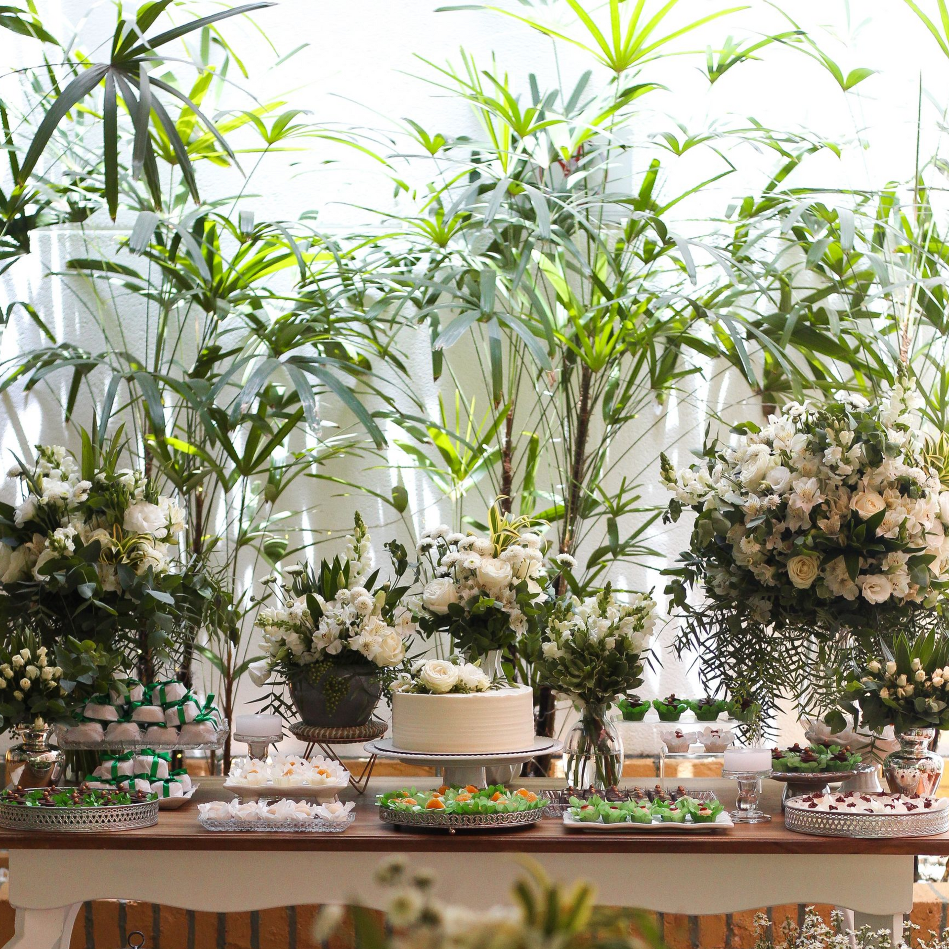Elegante mesa de arranjo floral com flores brancas, plantas verdes semelhantes a bambu e pequenos objetos decorativos.