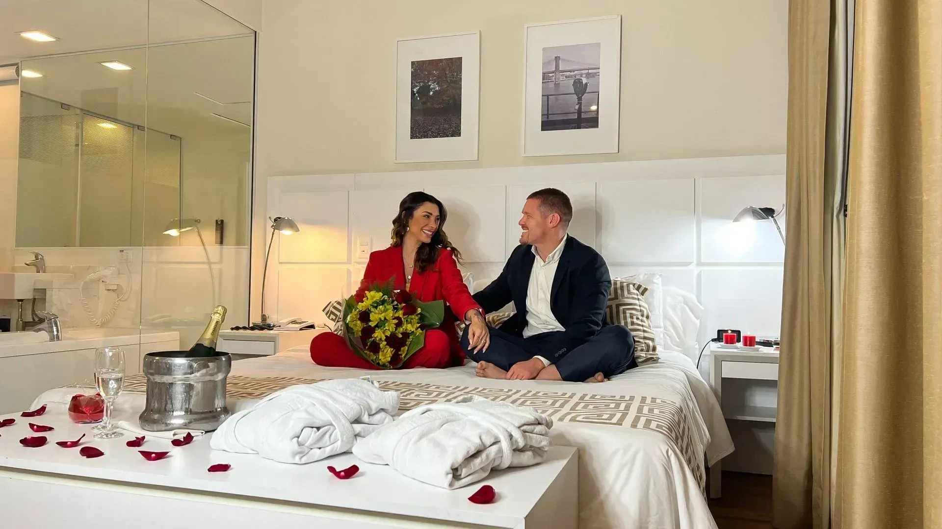 Um homem e uma mulher estão sentados em uma cama de mãos dadas, com buquê de rosas e garrafa de espumante.