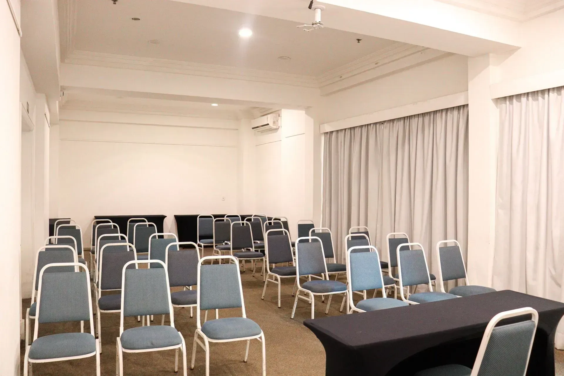 Sala de conferências vazia com fileiras de cadeiras azuis em frente a uma mesa coberta com uma toalha preta, sob luzes fortes no teto.