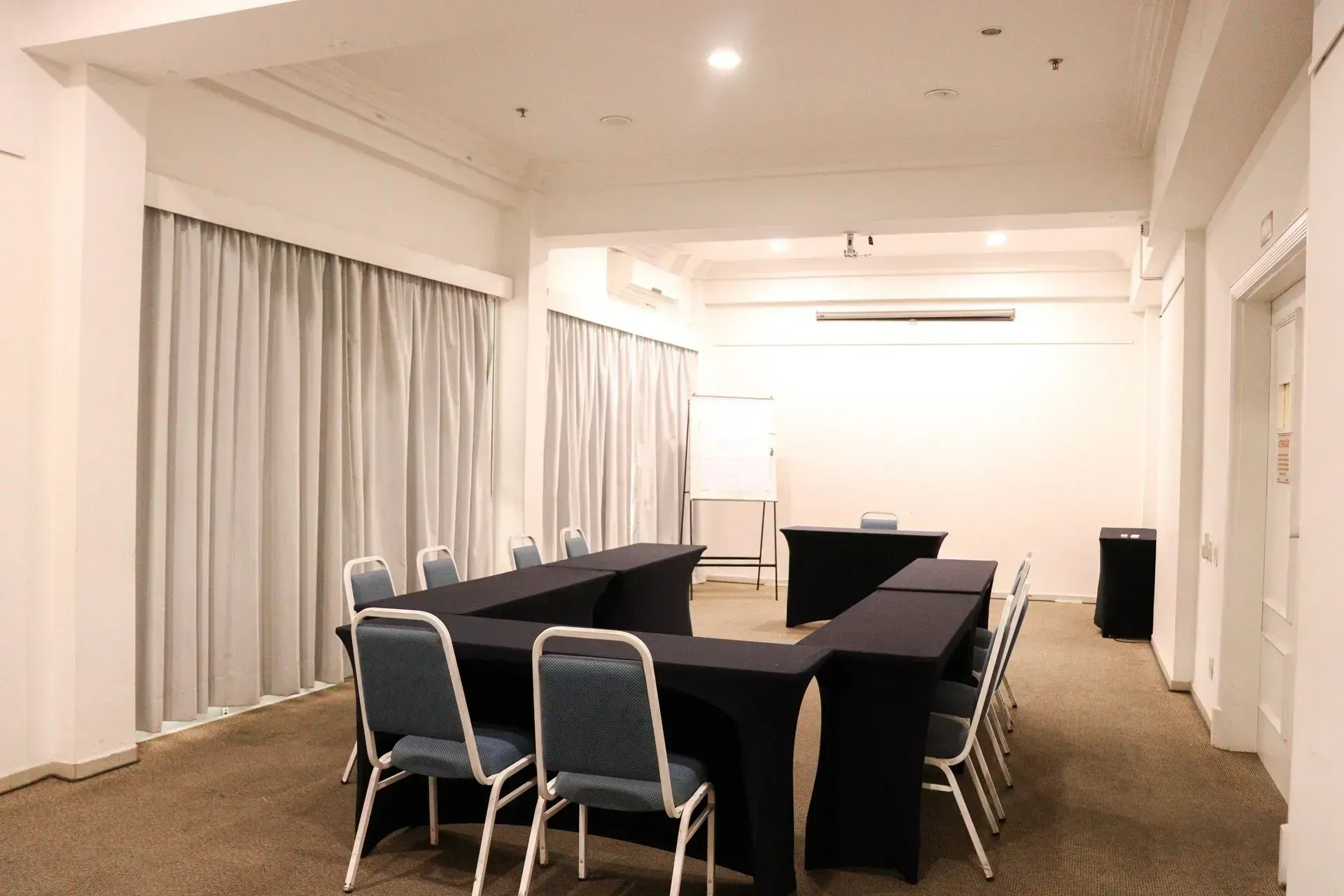 Sala de conferências vazia com mesas pretas, cadeiras cinzentas e cortinas fechadas sob luzes fortes no teto.
