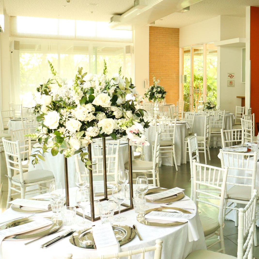 Elegante recepção de casamento em branco com arranjos florais e mesas postas em um salão iluminado.