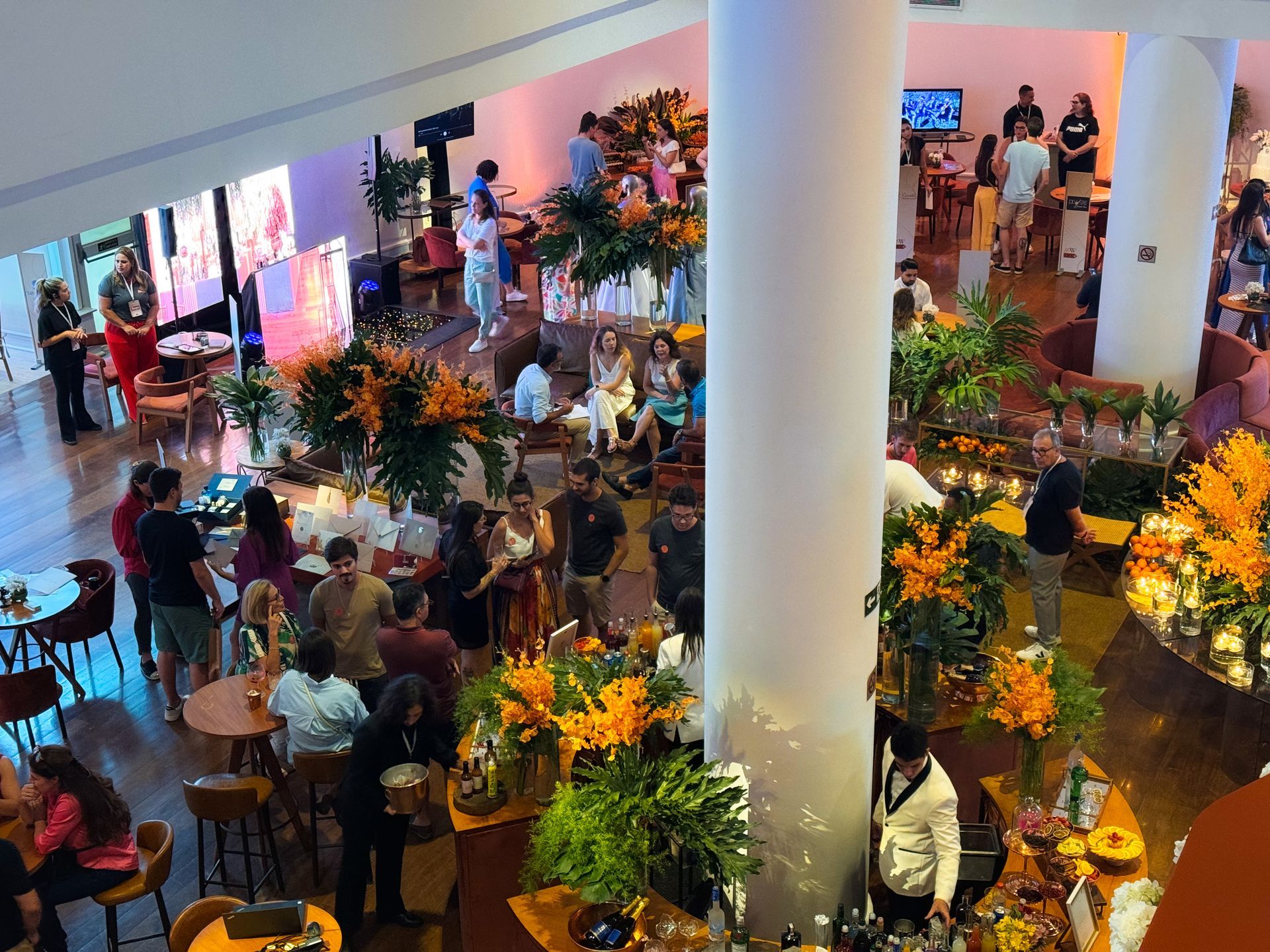 Evento lotado em ambiente fechado, com pessoas ao redor de arranjos florais e altas colunas brancas em um salão iluminado.