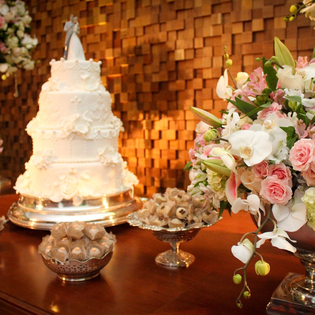 Bolo de casamento branco de vários andares com arranjos florais sobre uma mesa de madeira contra um fundo âmbar quente.