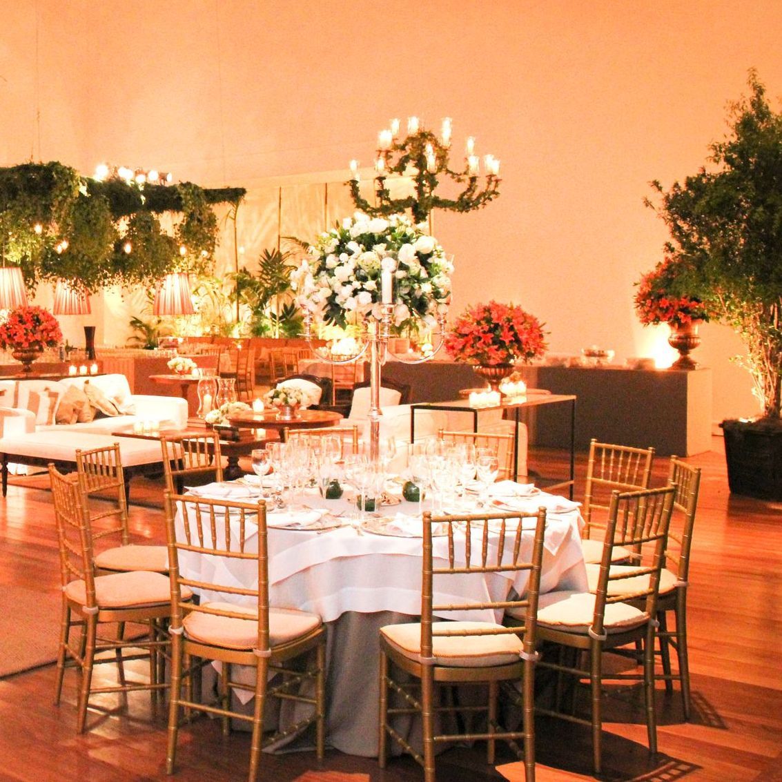 Elegante salão de banquetes com mesas redondas cobertas com toalhas brancas, cadeiras douradas, arranjos florais e iluminação aconchegante.