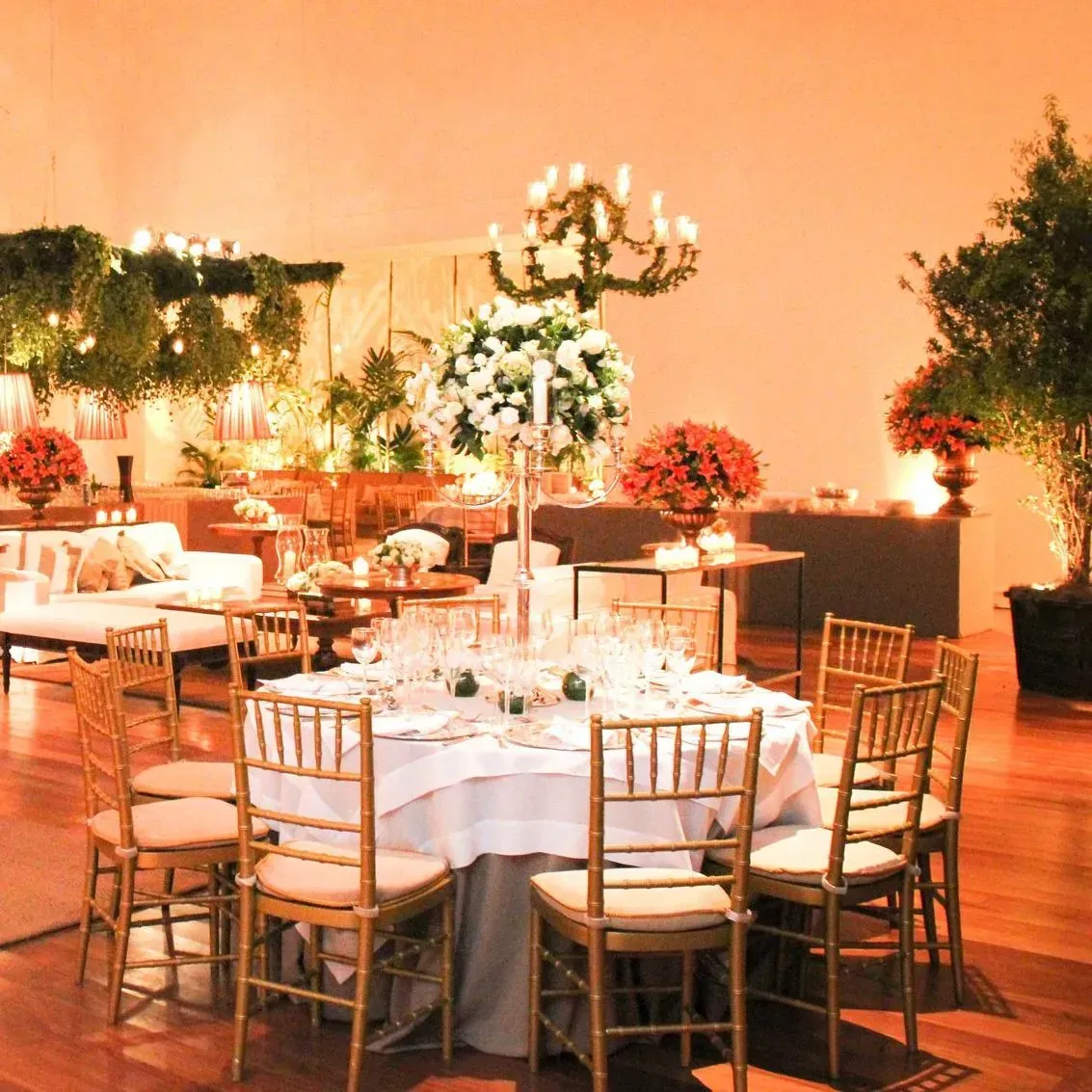 Elegante salão de banquetes com mesas redondas cobertas com toalhas brancas, cadeiras douradas, arranjos florais e iluminação aconchegante em tons de laranja.