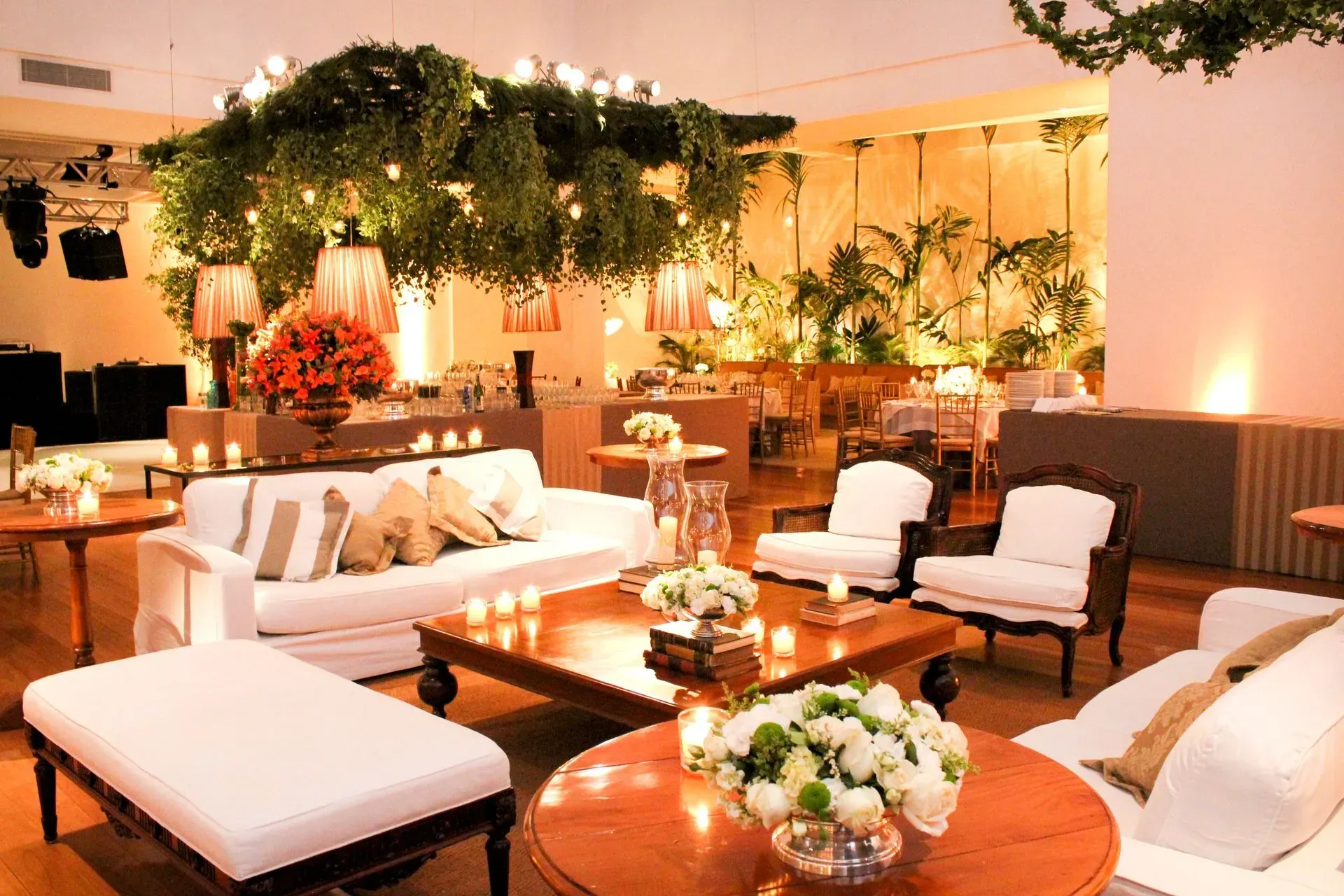 Sala de estar elegante com sofás brancos, mesas de madeira, iluminação aconchegante e plantas verdejantes.