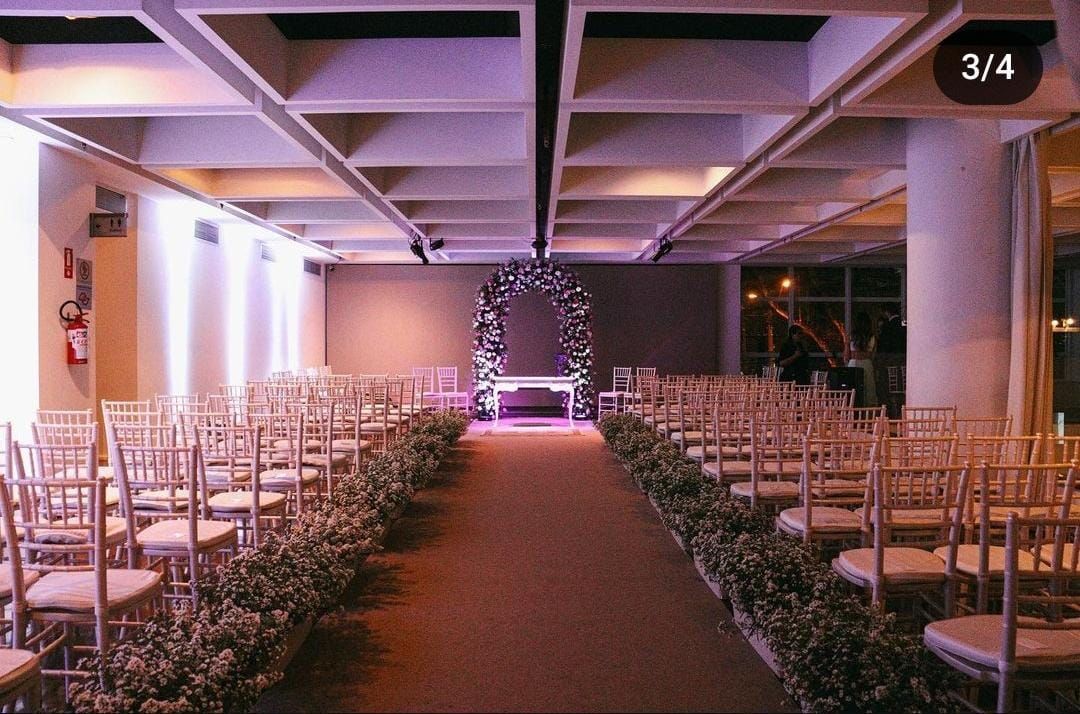 Um corredor de casamento ao ar livre com cadeiras brancas, decoração floral e um arco iluminado ao fundo.