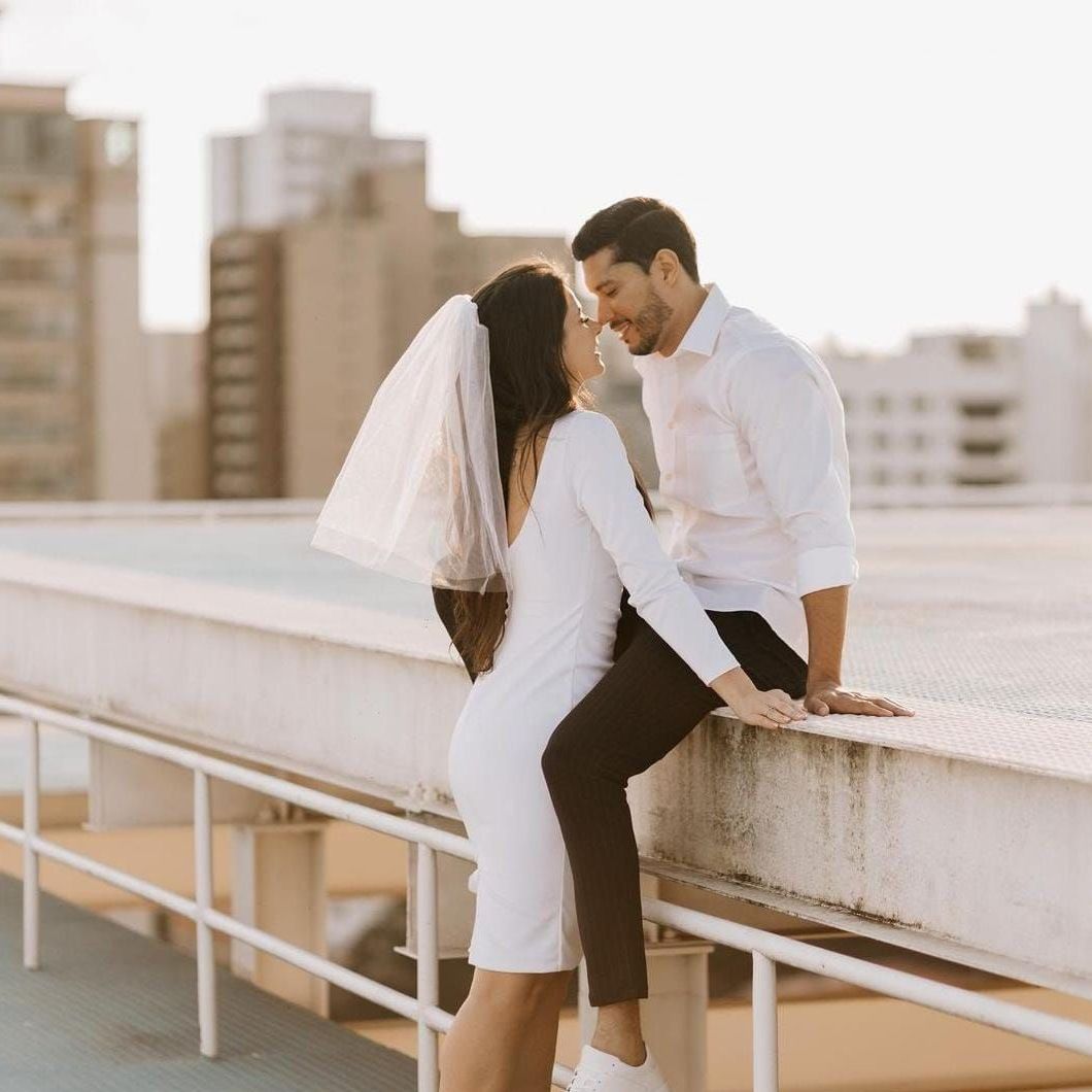 Casal vestido de branco se abraçando em um terraço com prédios da cidade ao fundo.