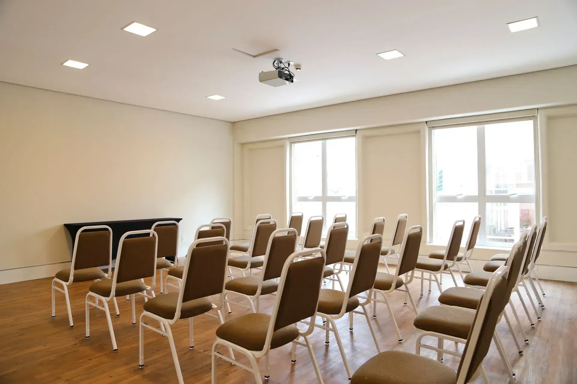 Sala de reuniões iluminada com fileiras de cadeiras vazias e uma tela de projeção perto de grandes janelas.