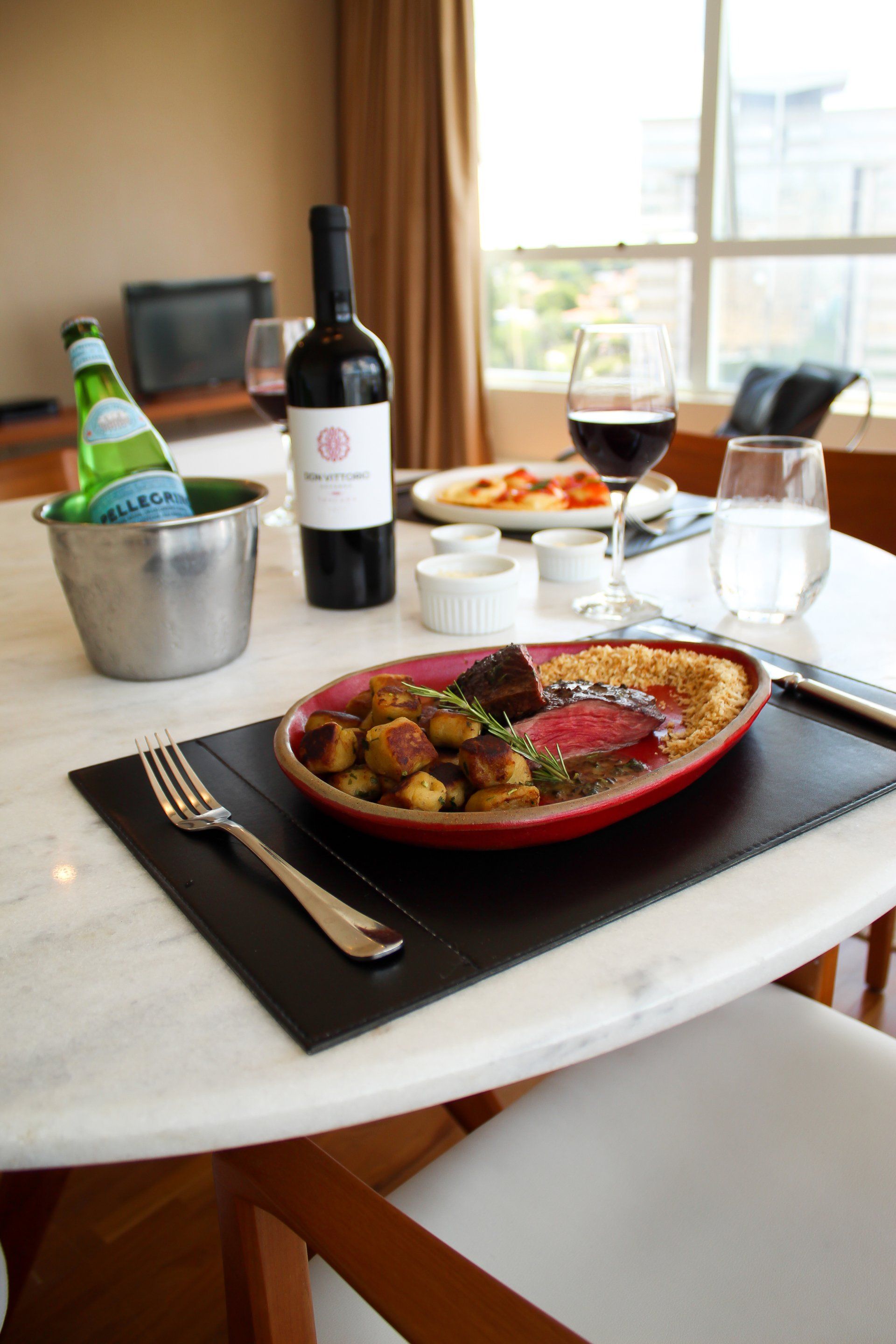 Prato vermelho com carne e legumes sobre uma mesa de jantar com vinho, água e vista para a janela.