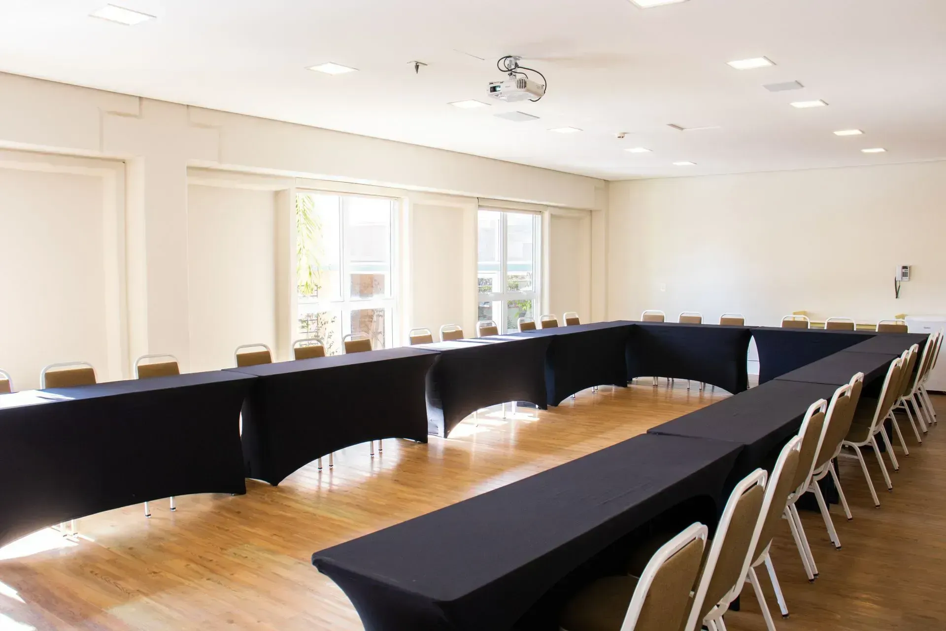 Sala de conferências vazia com mesas em formato de U, toalhas de mesa pretas e fileiras de cadeiras sobre um piso de madeira.