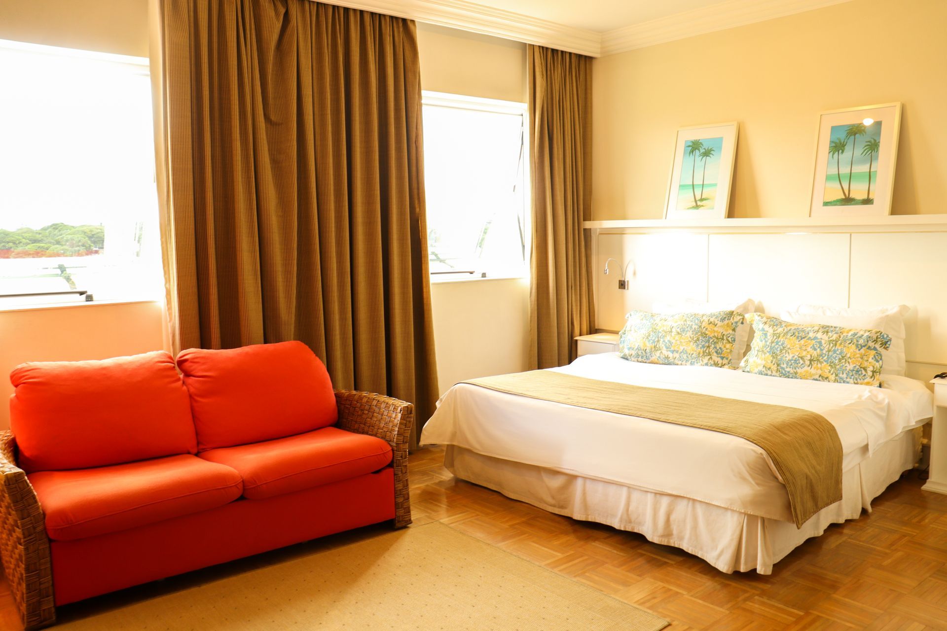 Quarto luminoso com cama branca, sofá laranja e cortinas grandes junto a janelas ensolaradas.