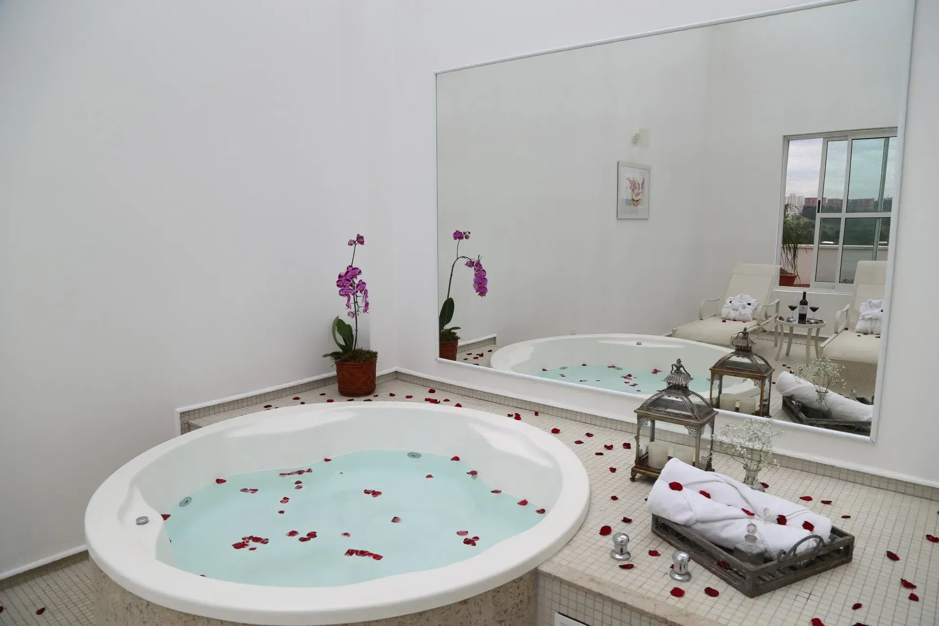 Banheiro estilo spa com banheira de imersão branca, pétalas de rosa, velas e toalhas dobradas ao lado de um espelho.