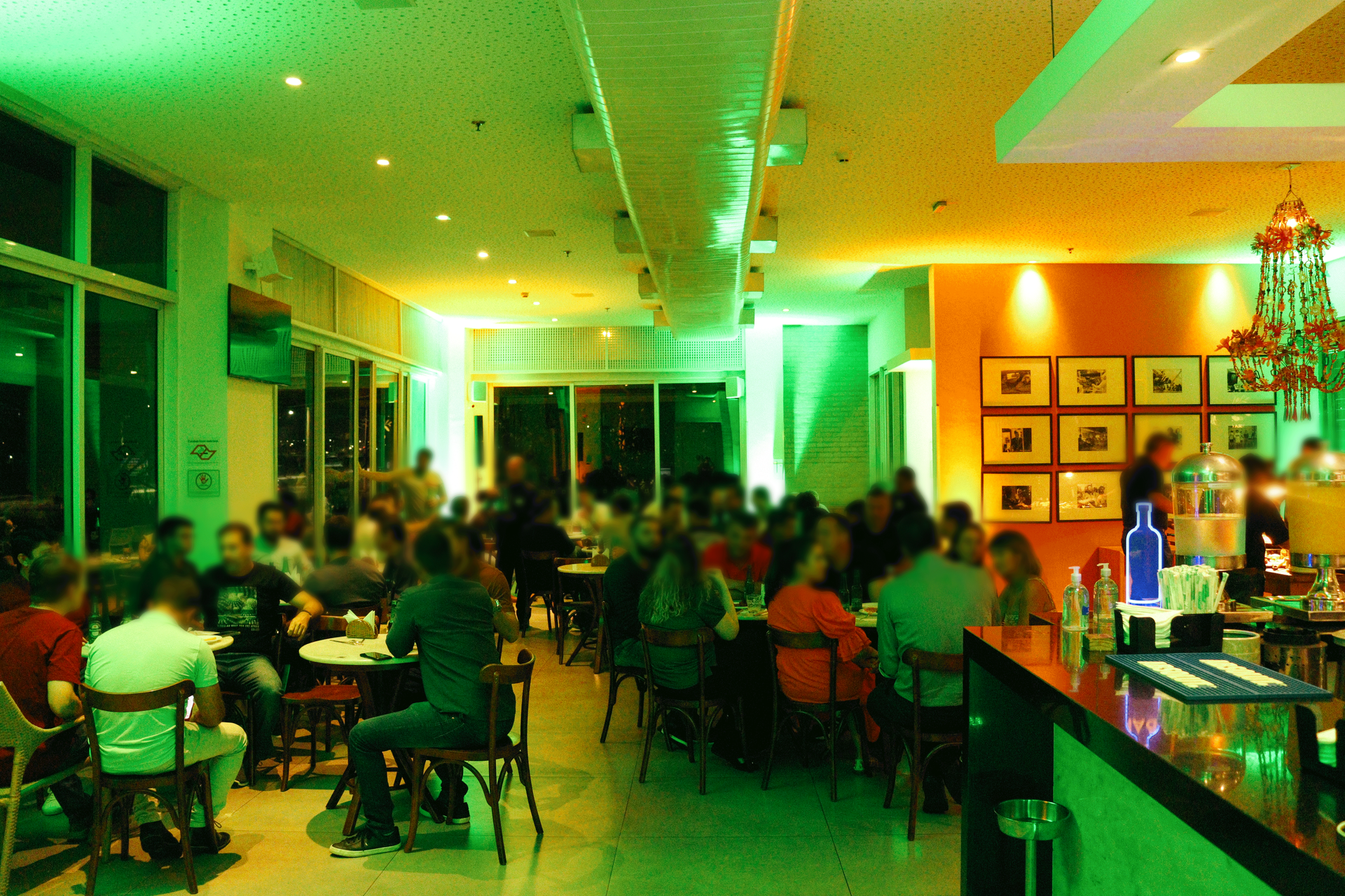 Interior de restaurante lotado, iluminado em verde, com clientes sentados às mesas e um bar à direita.