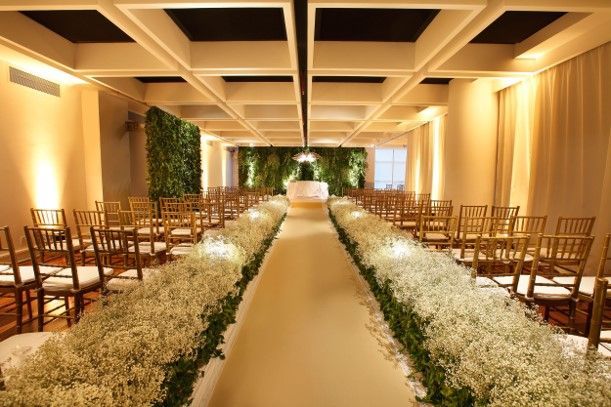 Elegante corredor de casamento interno com arranjos florais brancos e fileiras de cadeiras sob iluminação aconchegante.