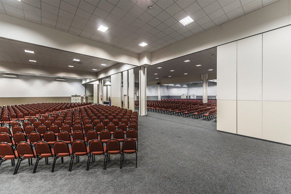Sala de conferências vazia com fileiras de cadeiras vermelhas, carpete cinza e teto claro.