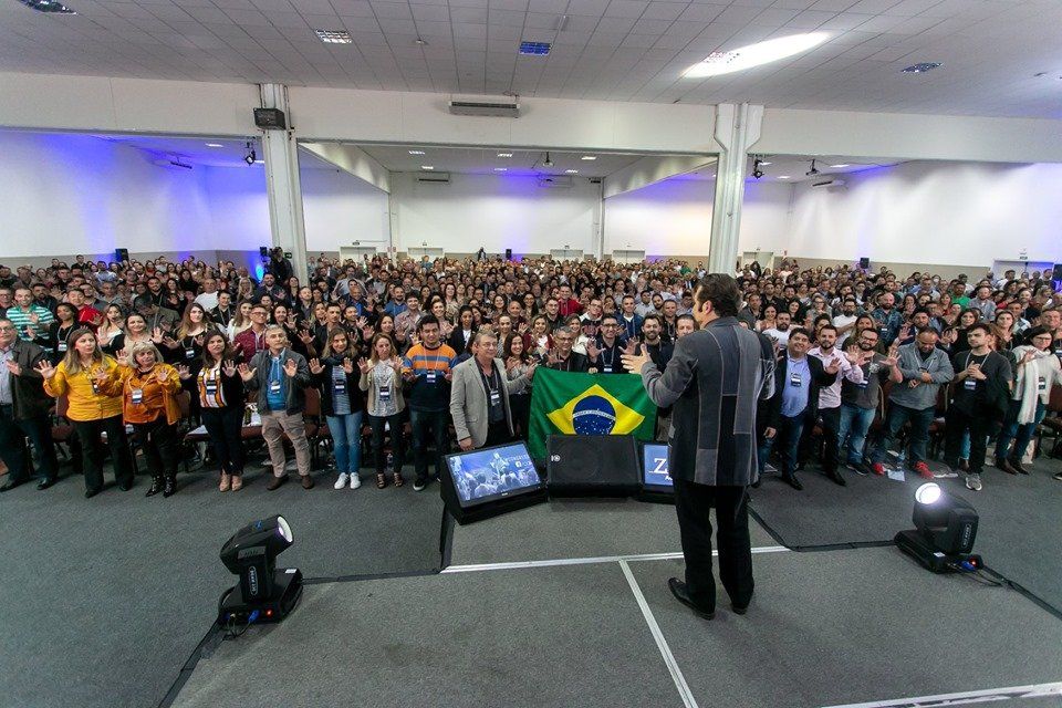 Palestrante de frente para uma grande plateia em um auditório, com a bandeira do Brasil e as luzes do palco visíveis.