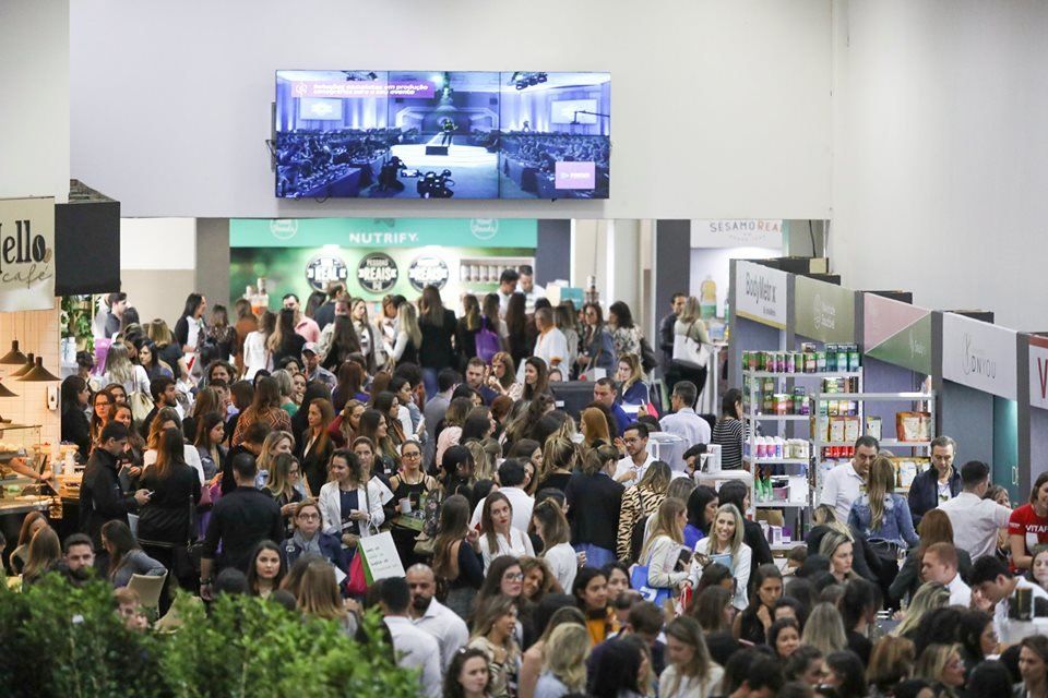 Salão de exposições interno lotado, com pessoas visitando os estandes e um grande telão digital acima.