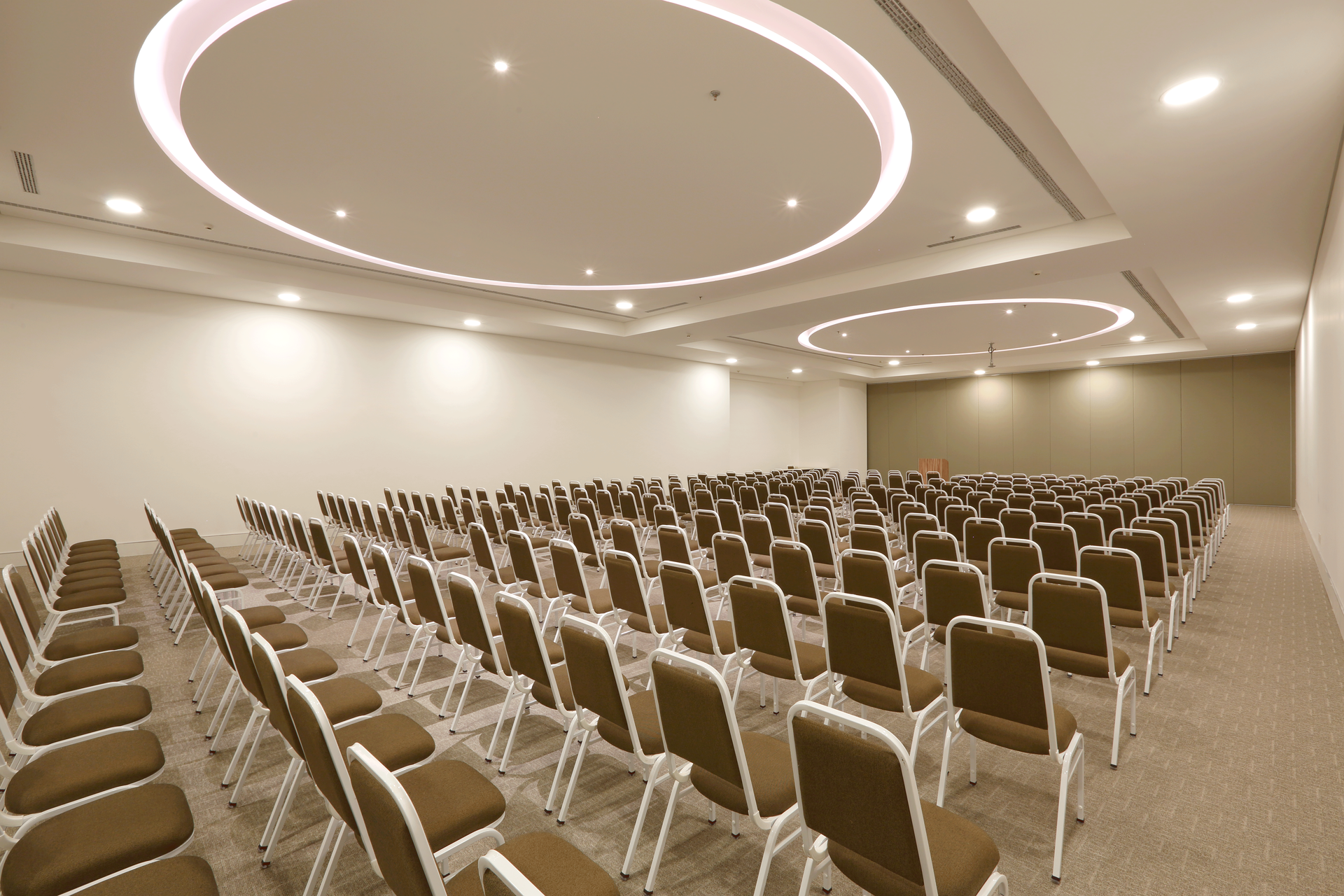 Sala de conferências vazia com fileiras de cadeiras bege e luminárias circulares modernas no teto.