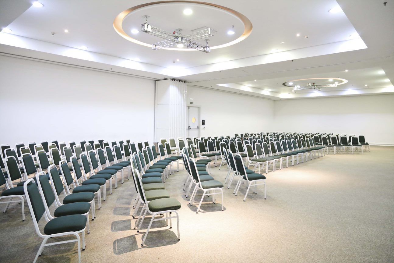 Sala de conferências vazia com fileiras de cadeiras verde-azuladas sob luzes brilhantes no teto.