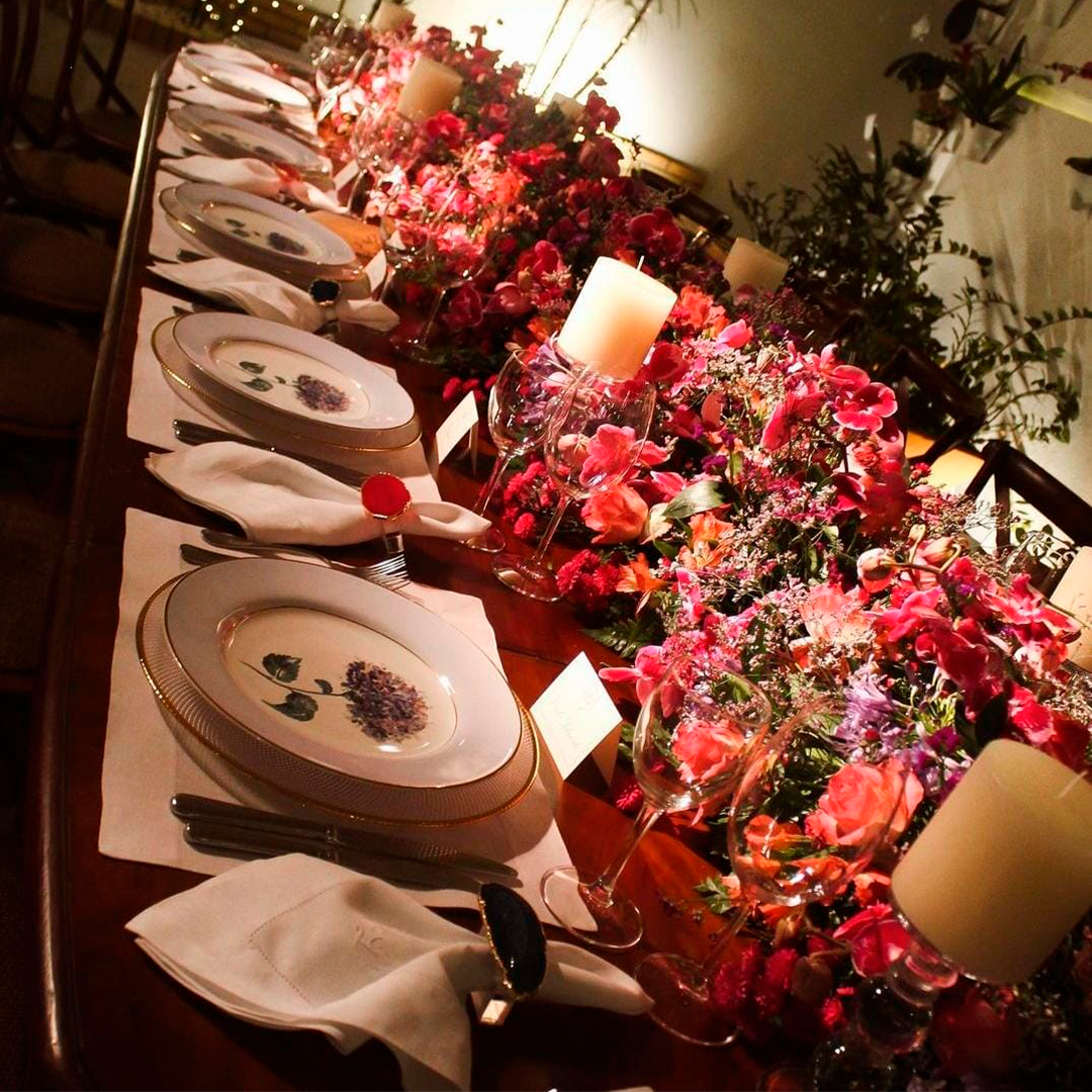 Mesa de banquete elegante com pratos brancos, velas e arranjos florais vermelhos sob iluminação aconchegante.