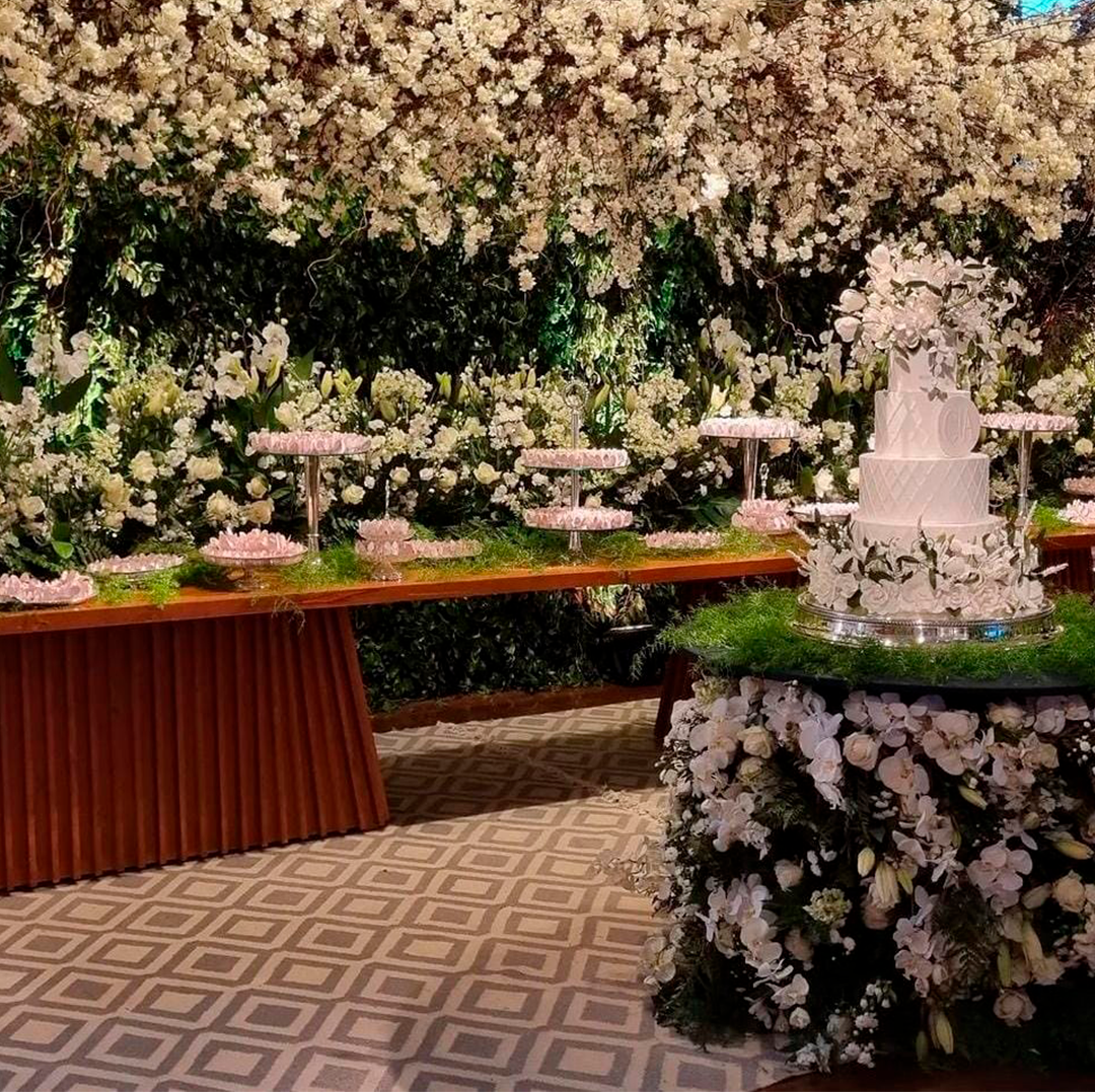Buffet de sobremesas coberto de flores com um bolo branco de vários andares e decoração floral rosa sobre um piso estampado.