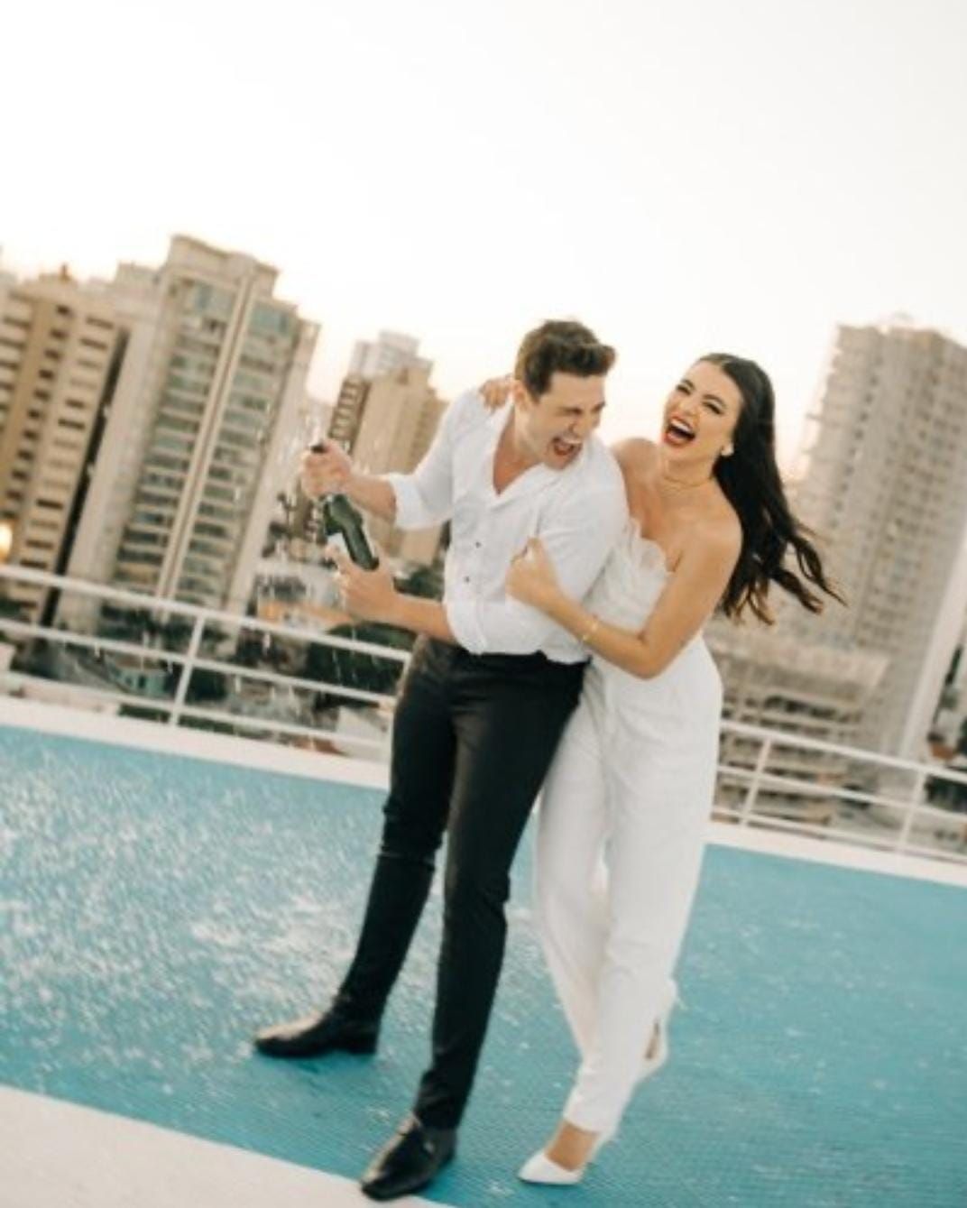 Casal vestido de branco rindo à beira de uma piscina na cobertura, com prédios da cidade ao fundo.