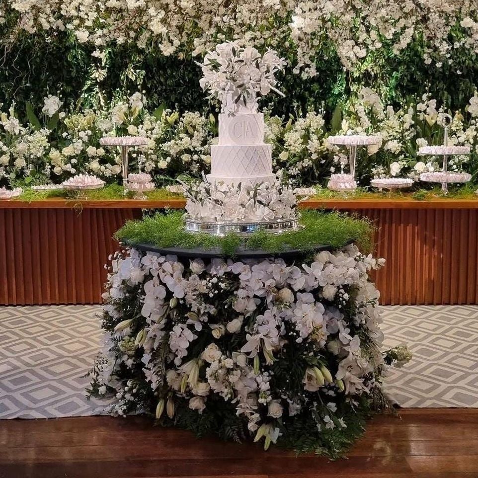 Bolo branco de vários andares sobre uma mesa coberta de flores em um salão de recepção decorado.